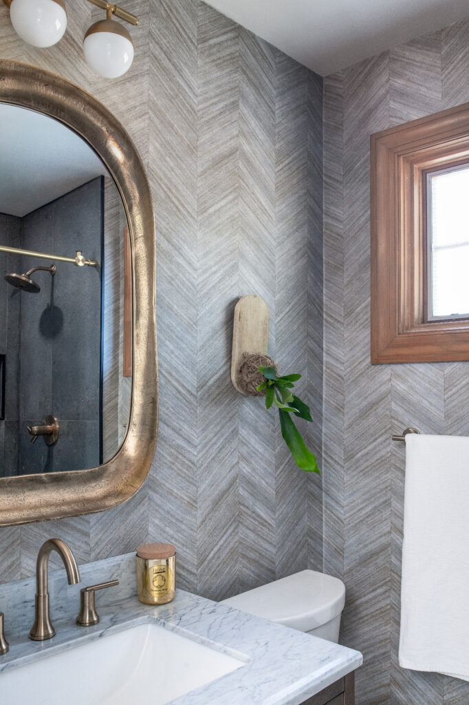 Looking into the corner, Plant mounted on wall Wallpaper on wall, Gold Oval Square Mirror, 3 globe light, dark vanity and gold plumbing fixture