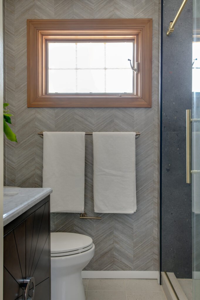 Looking toward the master bathroom window after renovation. Wallpaper on the wall, shower door and dark wood vanity